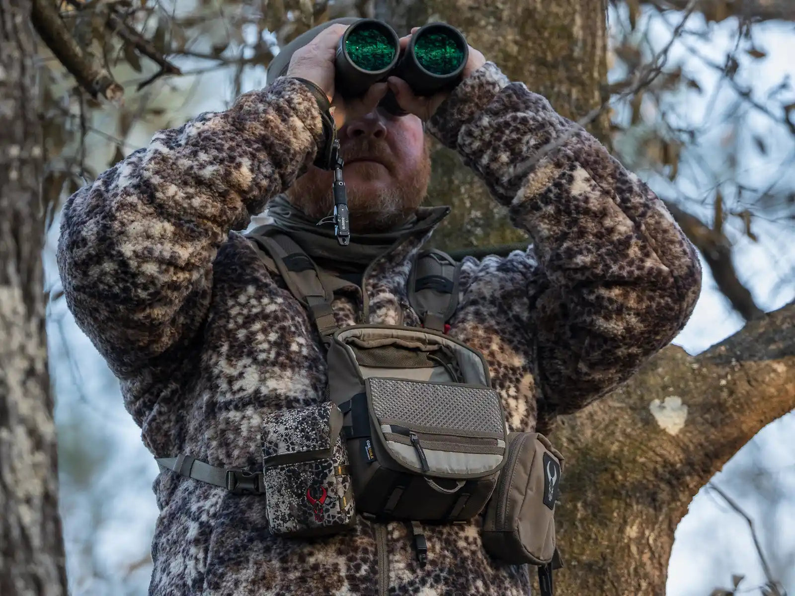 Person in camo fleece uses binoculars outdoors, wearing a Moonrivers chest pack with visible skull logo and gear pouches.