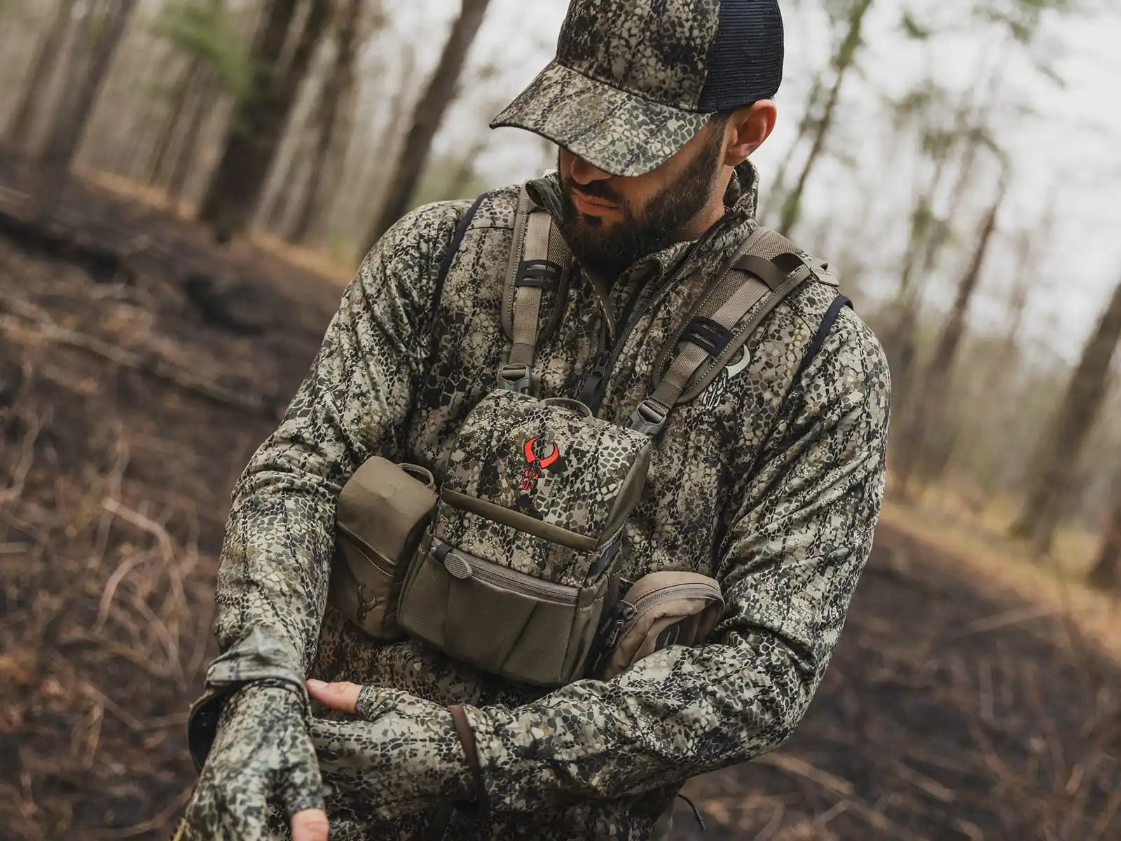 Person in camo gear and cap, wearing a Moonrivers chest pack with skull logo, standing in a forested area.