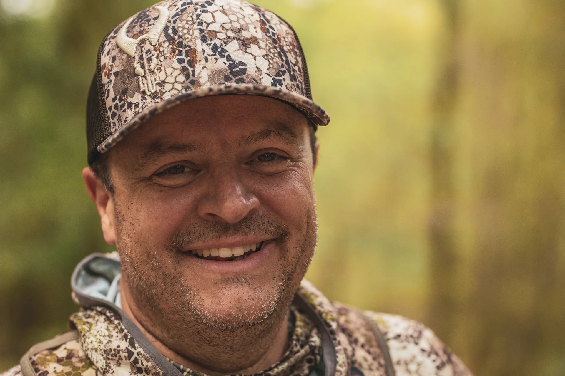 Person wearing Moonrivers camo hat with skull logo and matching camo jacket, outdoors in a forest setting.