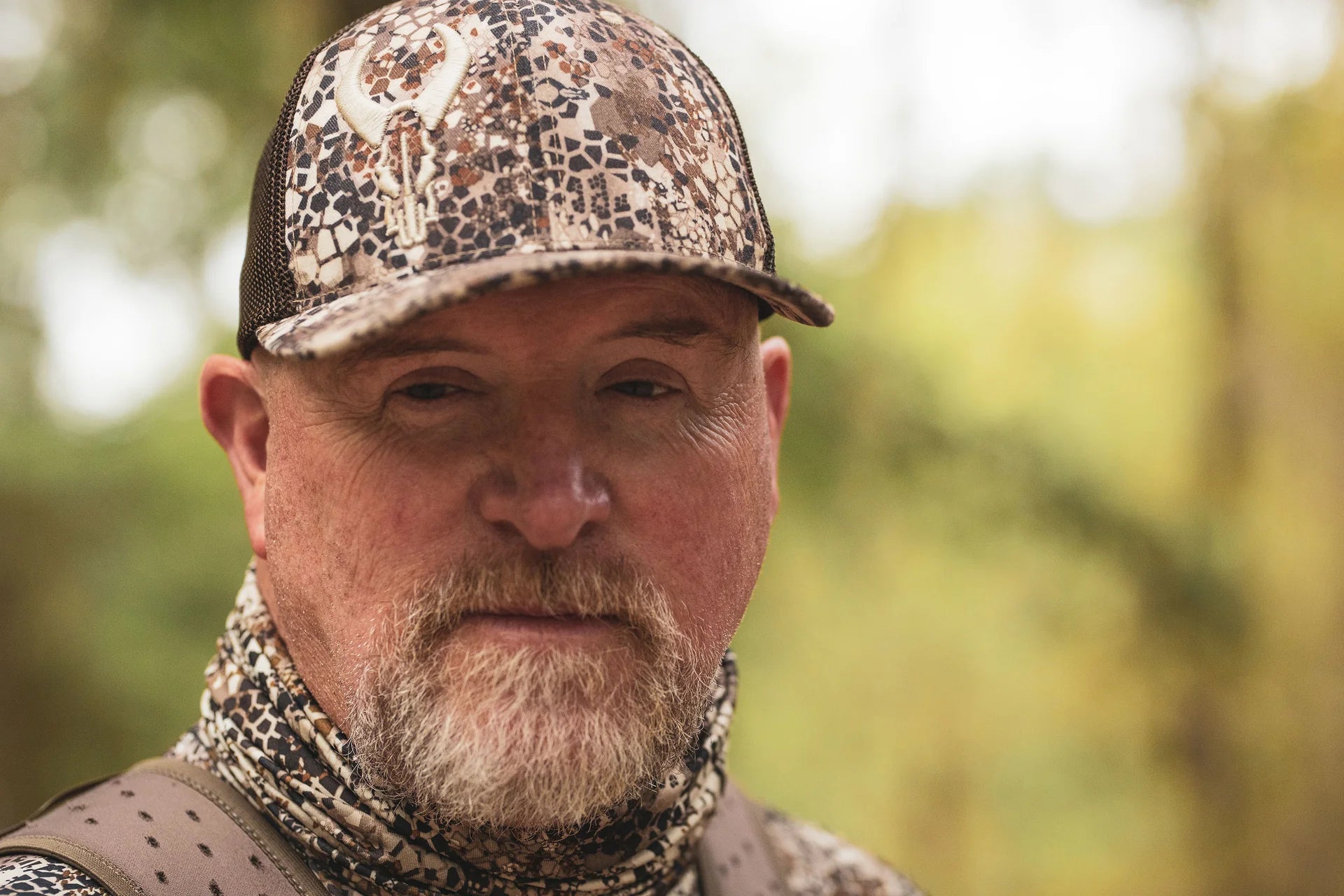 Person wearing Moonrivers camo hat with skull logo, matching neck gaiter, and backpack in an outdoor forest setting.