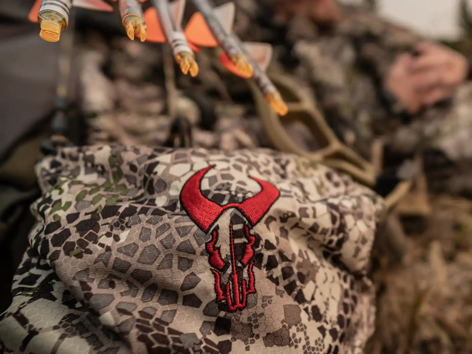 Close-up of a Moonrivers camo backpack with red skull logo, arrows above, and blurred hunter in background.