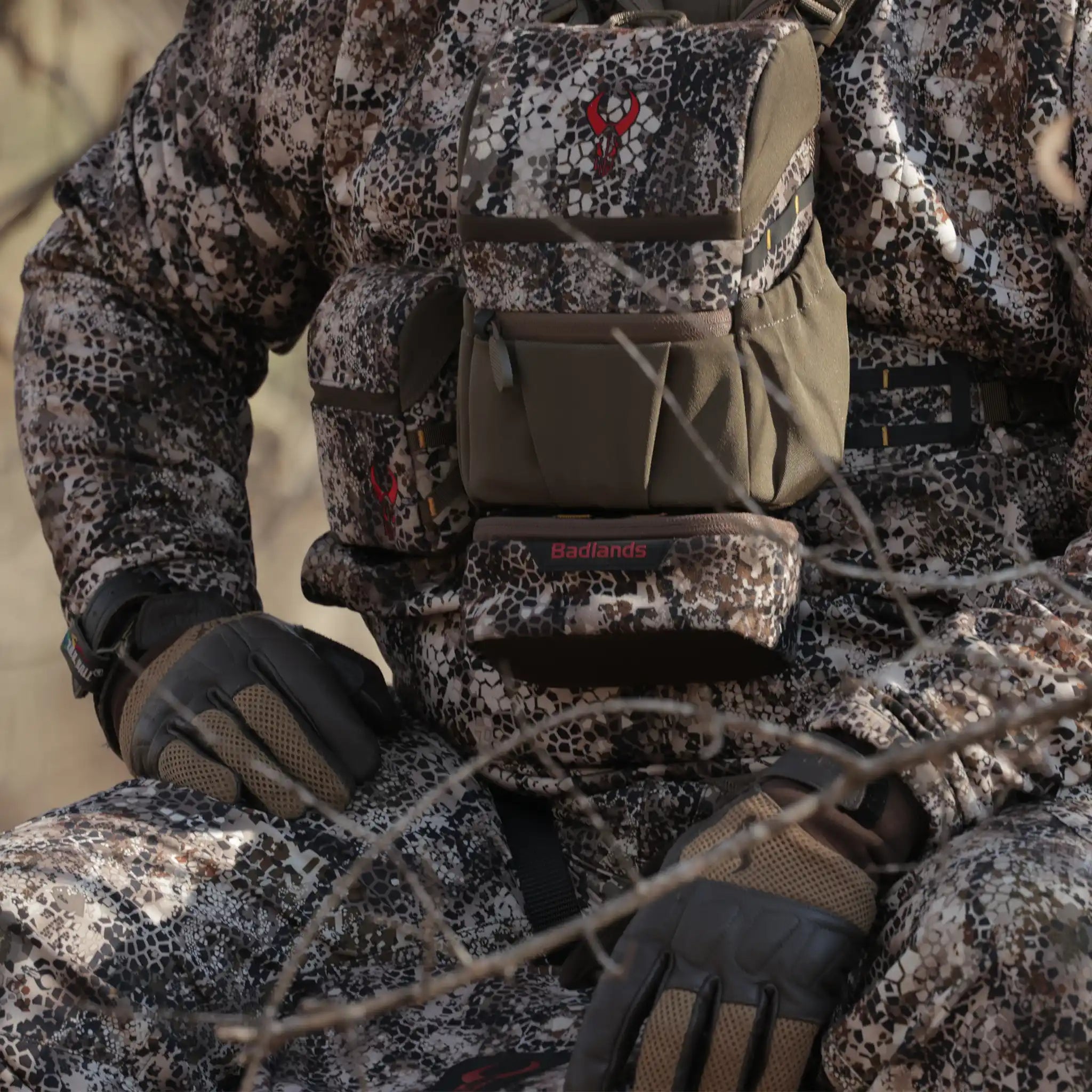 Person in camo gear with Moonrivers Logo on chest pack, sitting outdoors. Gloves and branches visible in foreground.