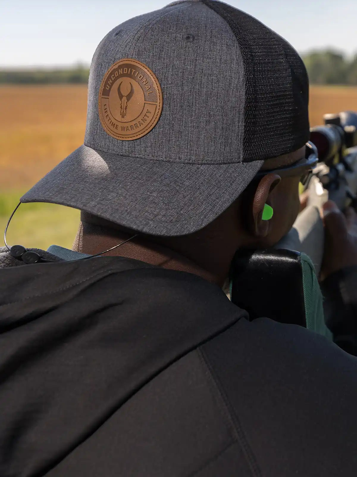 Person aiming a rifle outdoors, wearing a gray Moonrivers cap with skull logo and "Unconditional Lifetime Warranty" patch.