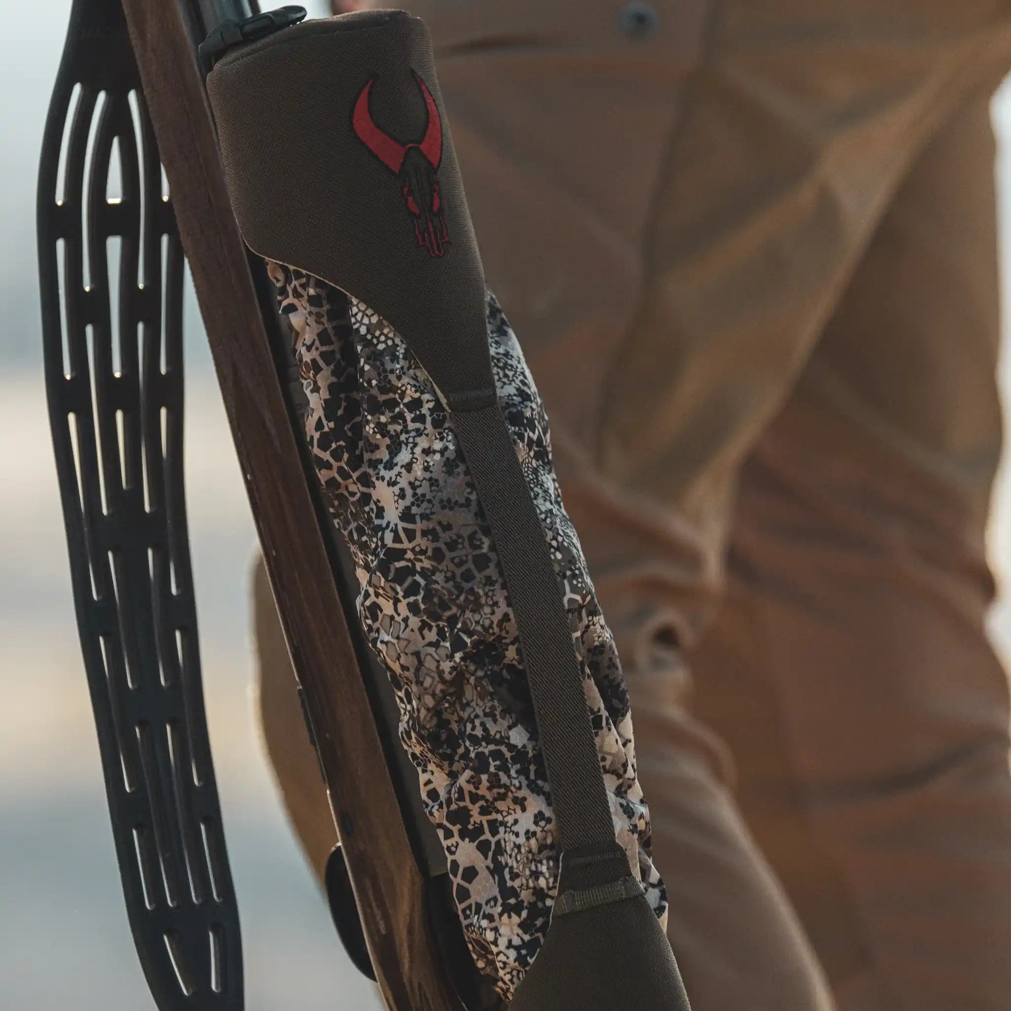 A rifle in a camo-patterned Moonrivers case with a red skull logo, held by a person in brown pants outdoors.