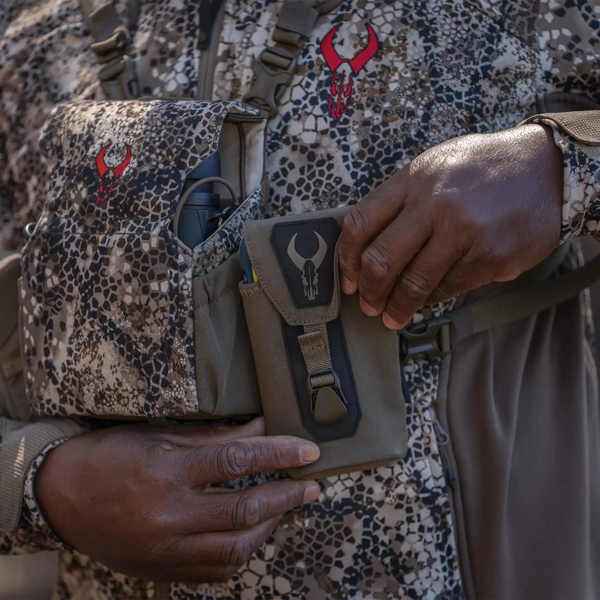 Person wearing camo gear with Moonrivers Logo, holding a pouch with the same skull-shaped Moonrivers Logo, outdoors.