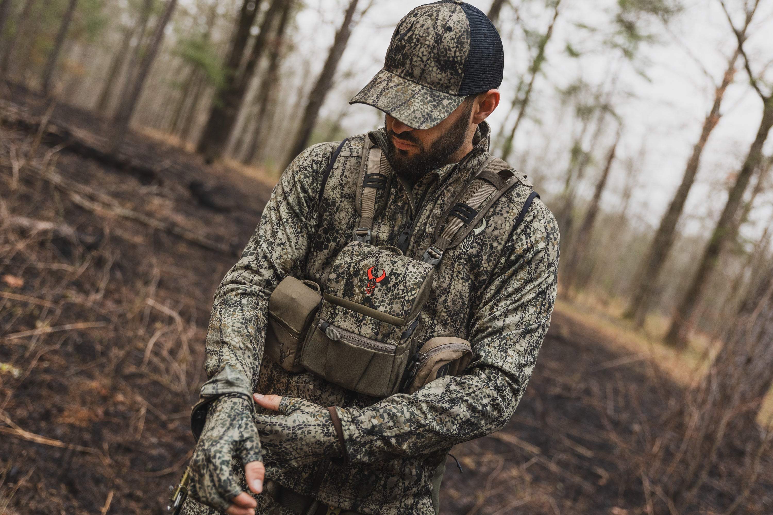 Person in camouflage gear and gloves outdoors, wearing a chest pack with the Moonrivers Logo in a wooded area.
