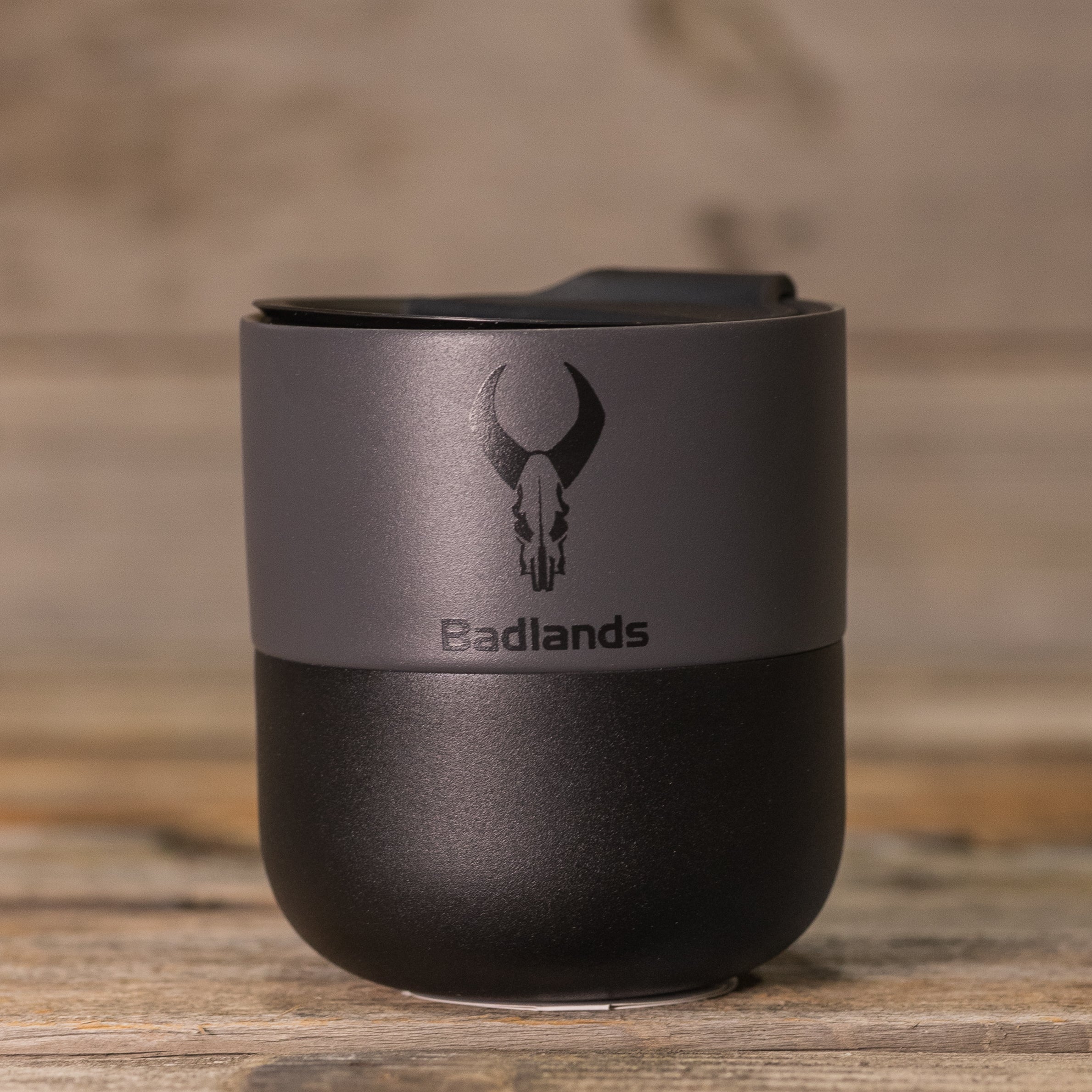 Black and gray travel mug with Moonrivers Logo (skull) and "Badlands" text, placed on a wooden surface.