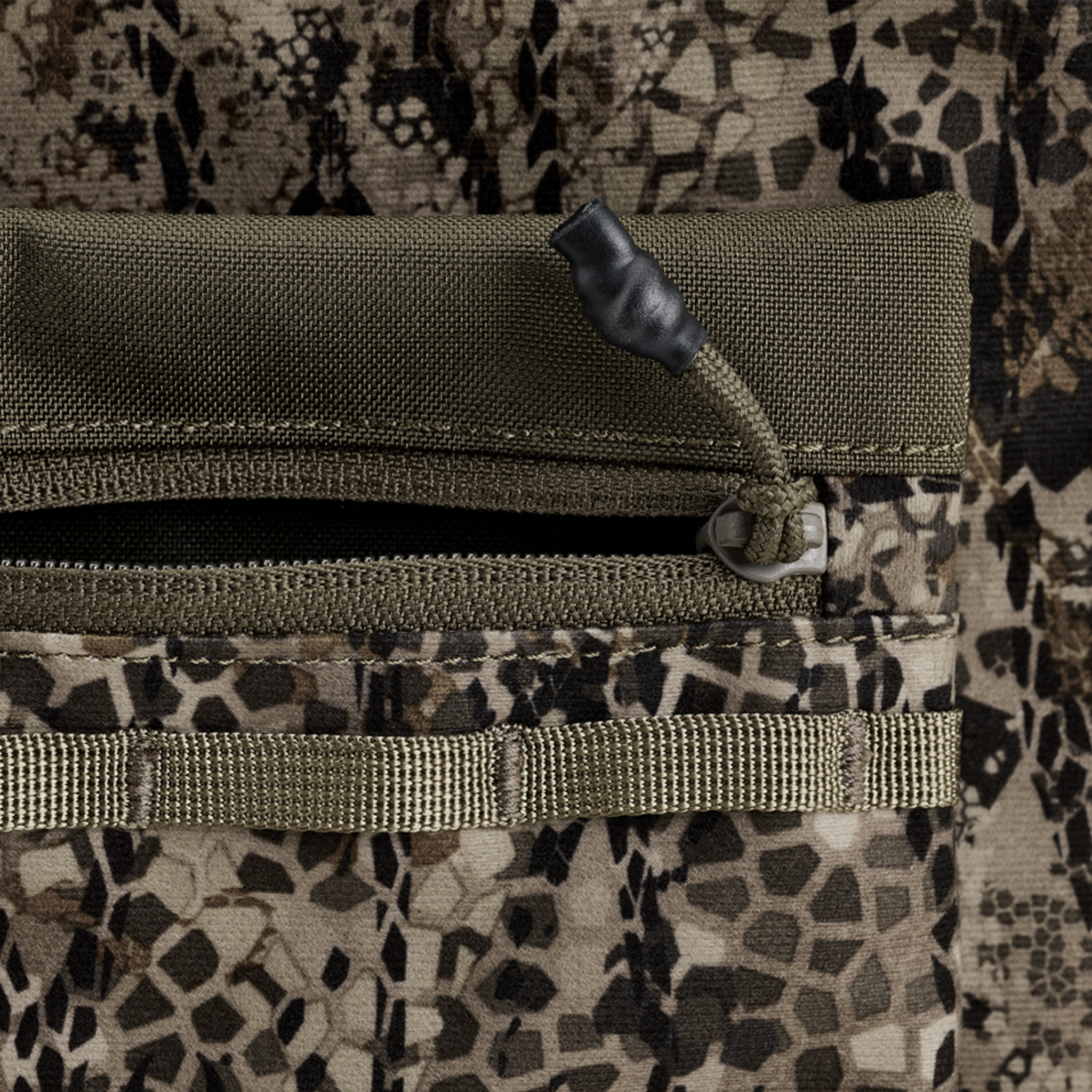 Close-up of a camouflage fabric pocket with a zipper and pull cord, featuring a textured pattern and webbing detail.