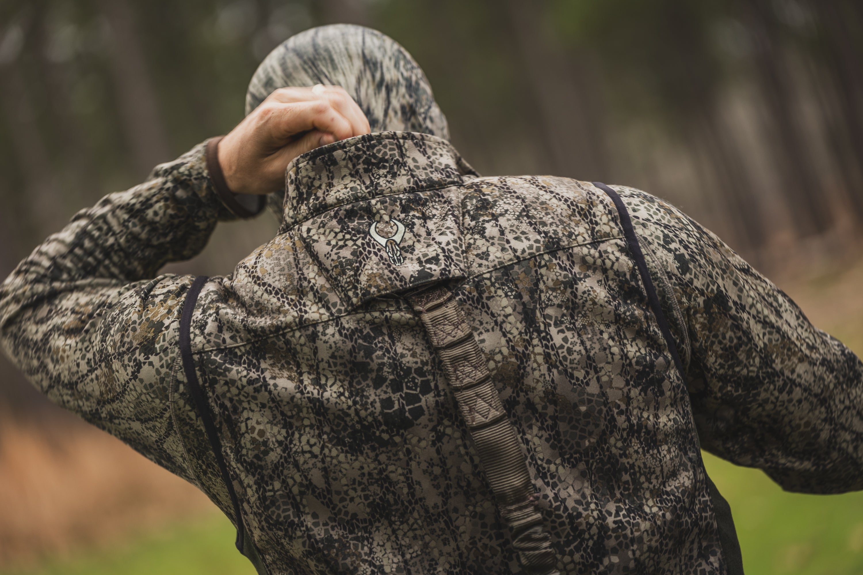Person in camo jacket outdoors, showing the Moonrivers skull logo on the back collar. Forest background, ADA-optimized.