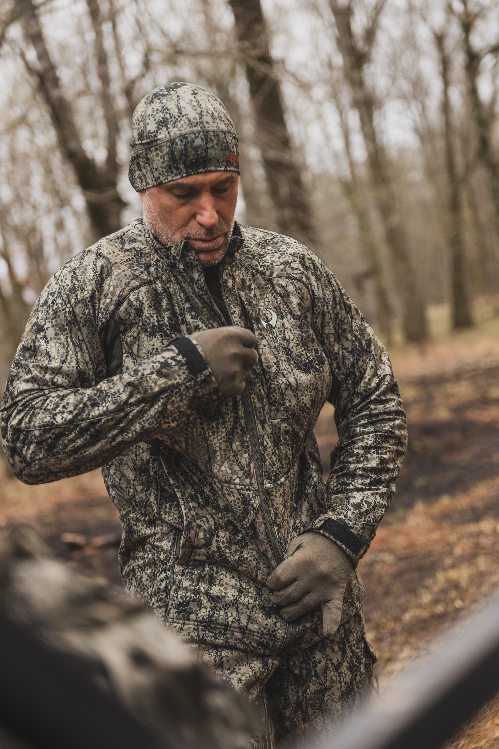Person in camo gear zipping jacket with Moonrivers Logo, standing outdoors among trees in a forest setting.