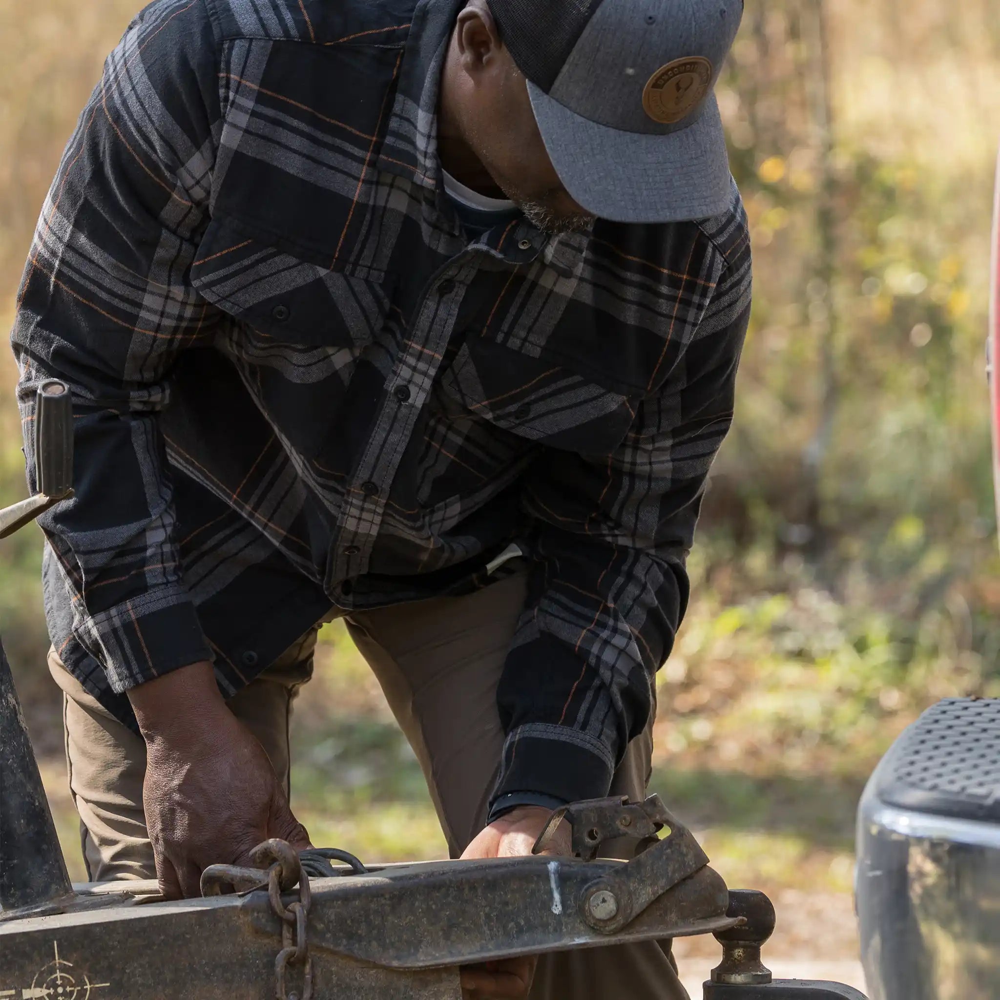 Man in plaid shirt and Moonrivers logo hat secures trailer hitch outdoors; autumn background.