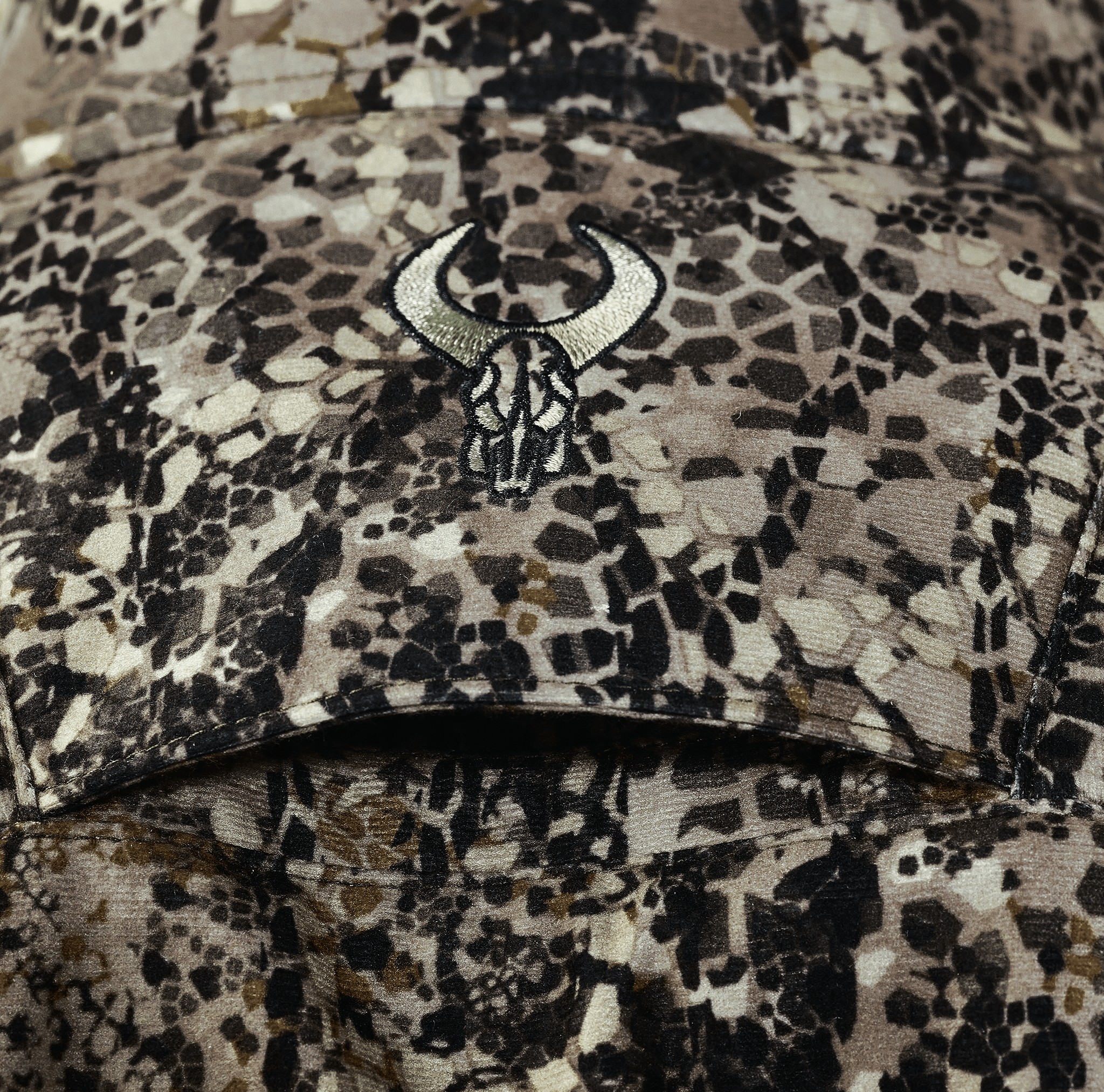Close-up of camo fabric with embroidered Moonrivers skull logo, featuring earth-tone geometric patterns for outdoor use.