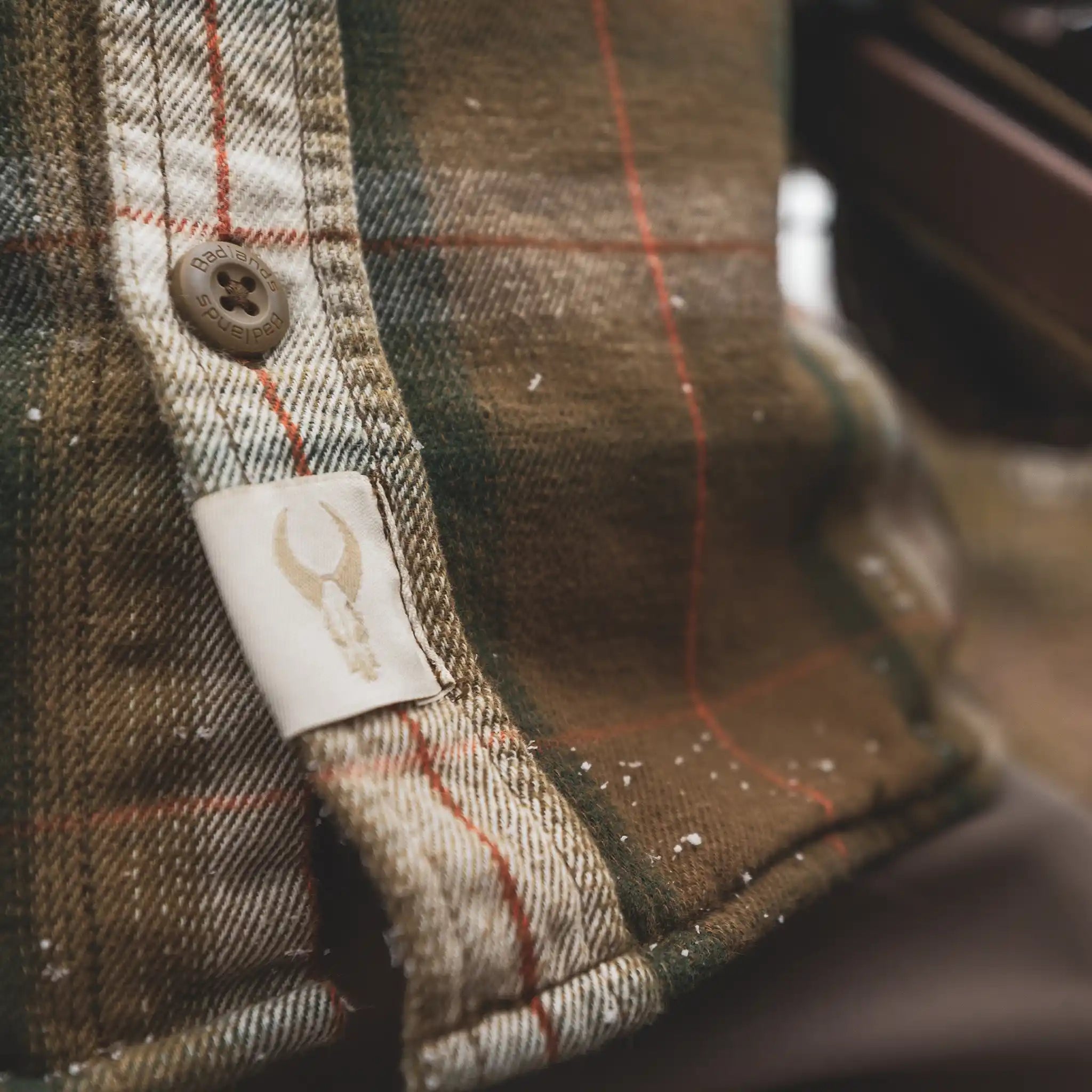 Close-up of a plaid Moonrivers shirt with a skull logo tag and branded button, dusted with light snow.