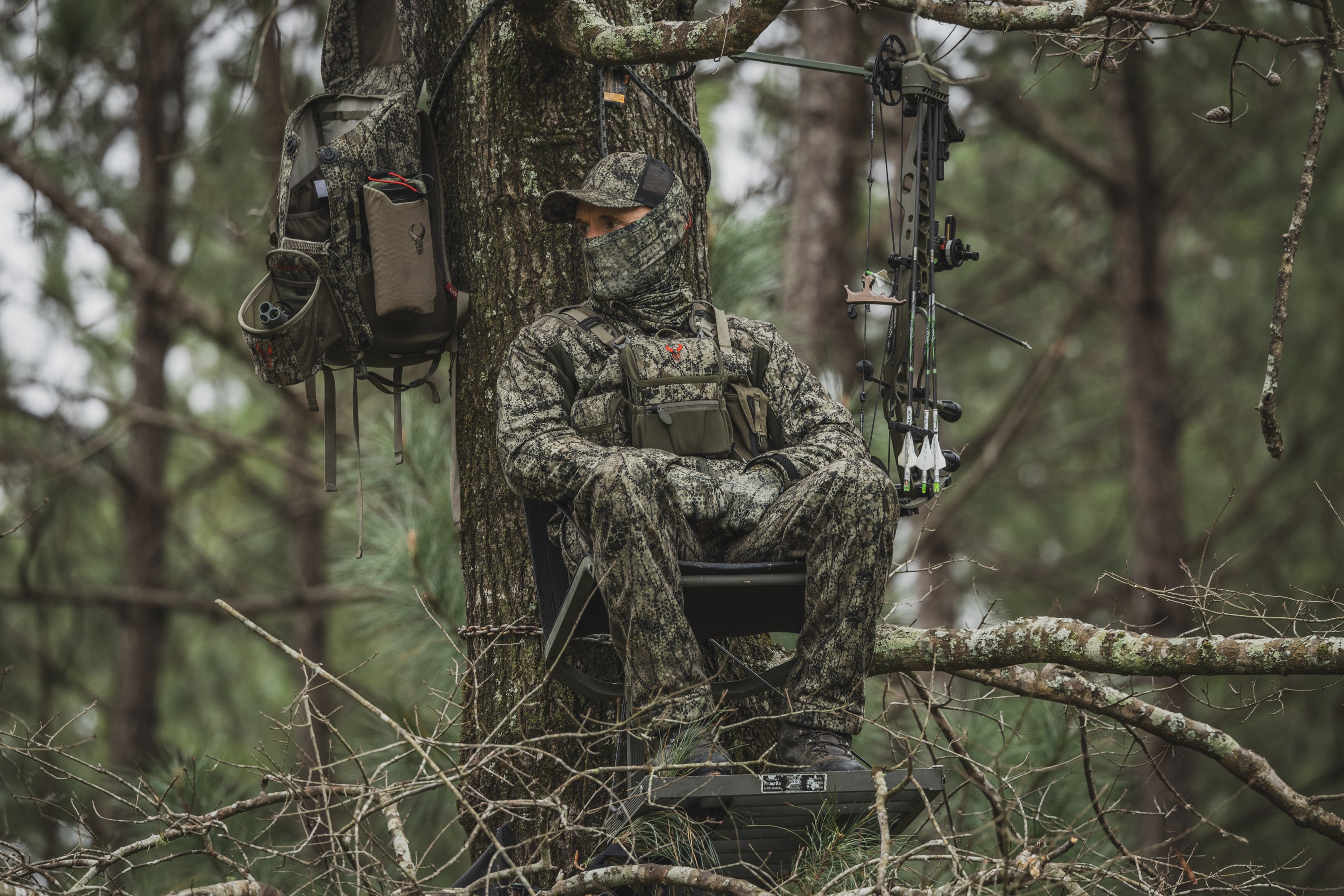 Hunter in camouflage sits in a tree stand with a bow; Moonrivers Logo visible on backpack and chest pouch.