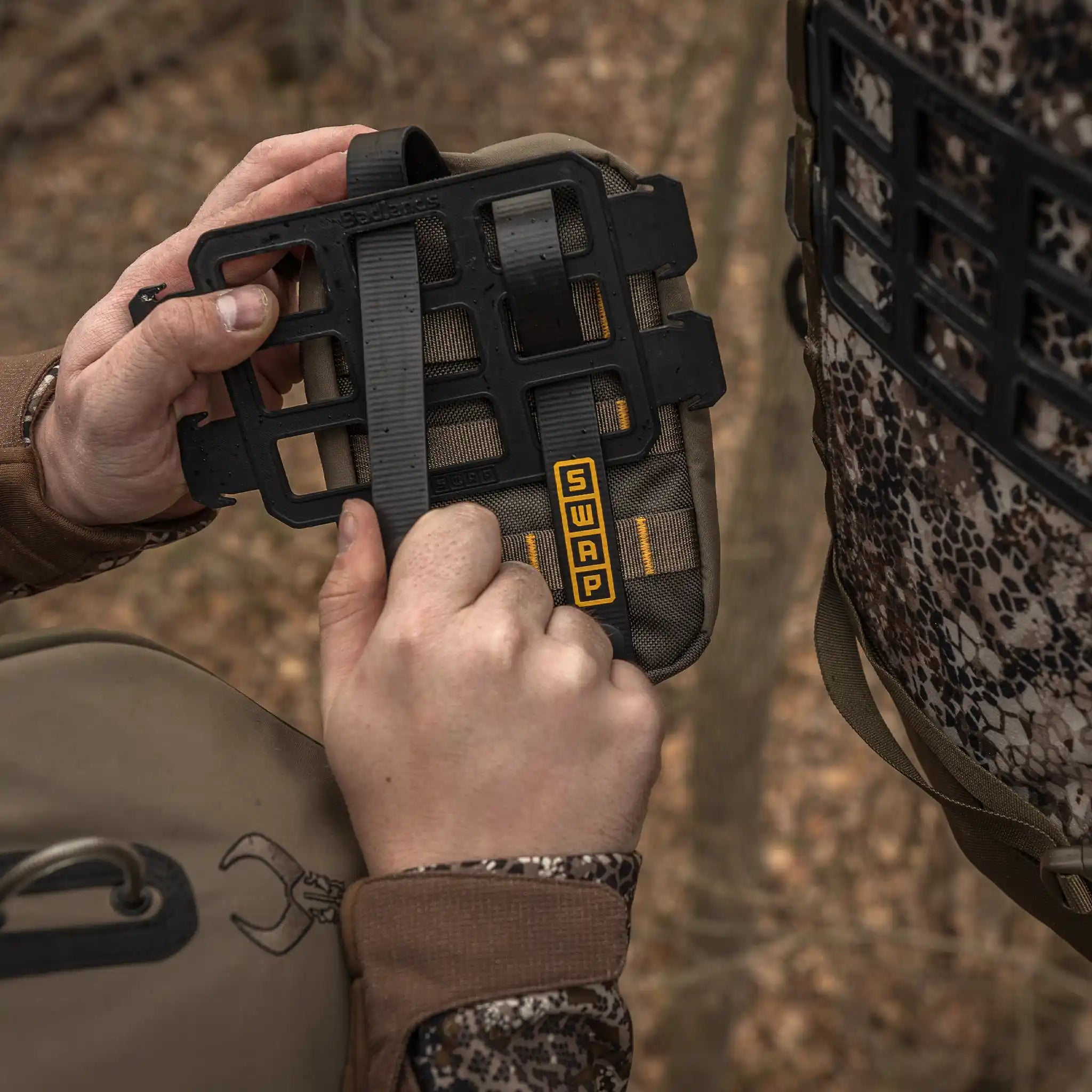 Hands attach a SWAP panel to a Moonrivers backpack using a modular grid system; outdoors, camo gear visible.