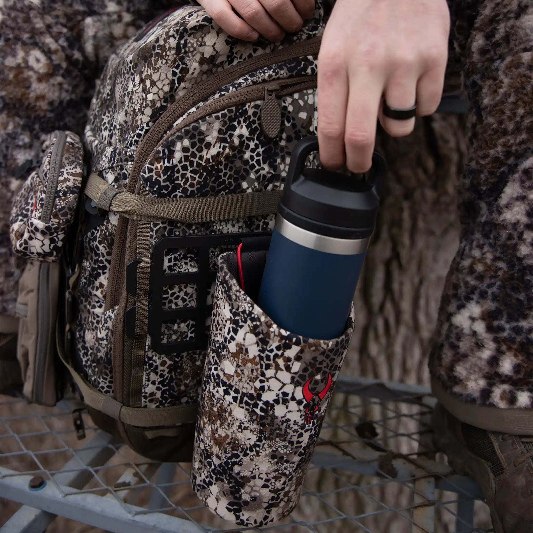Hand places a blue water bottle into a camo Moonrivers Logo pouch attached to a camo backpack outdoors.