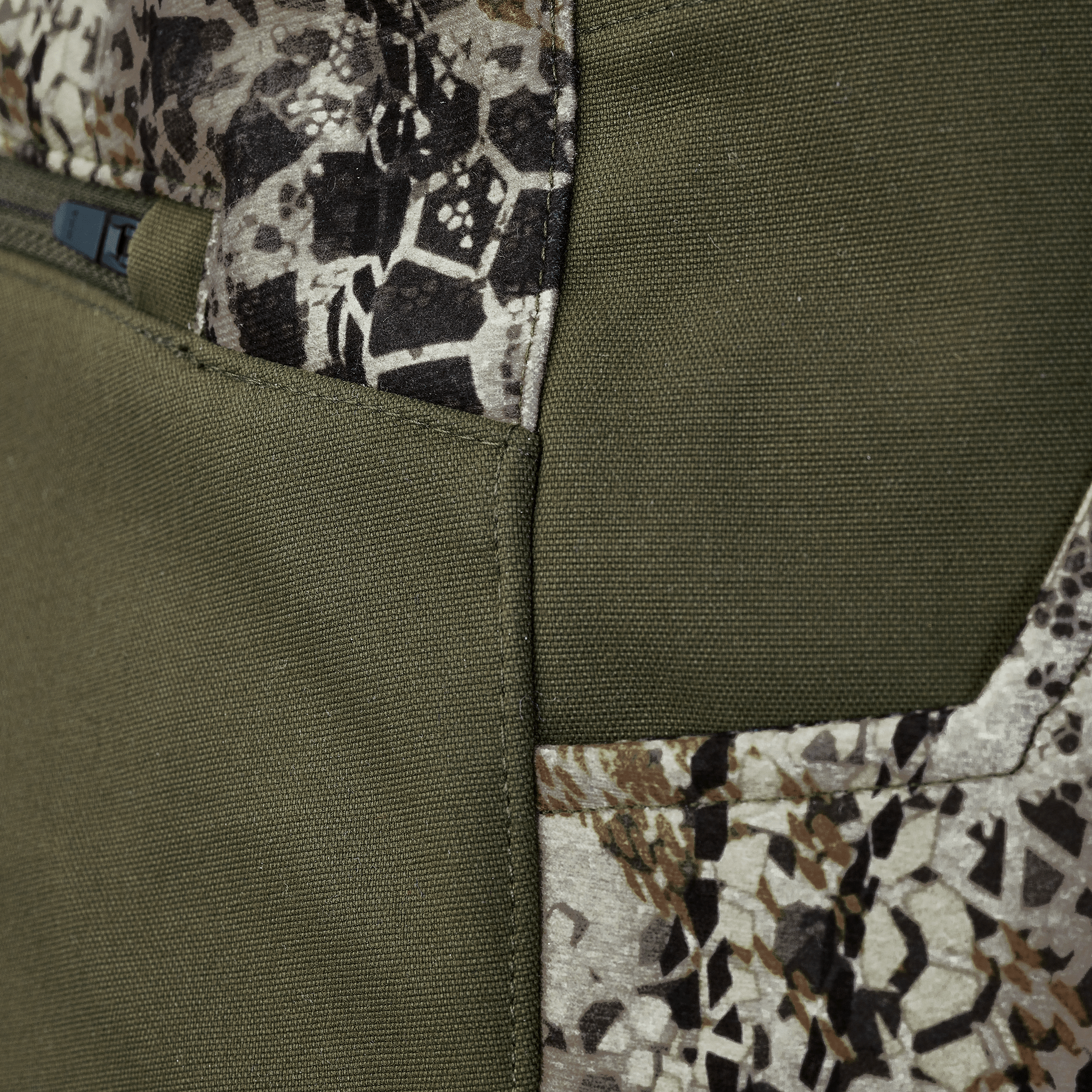 Close-up of a fabric with olive green and camo pattern, showing a seam and zipper. No Moonrivers Logo visible.