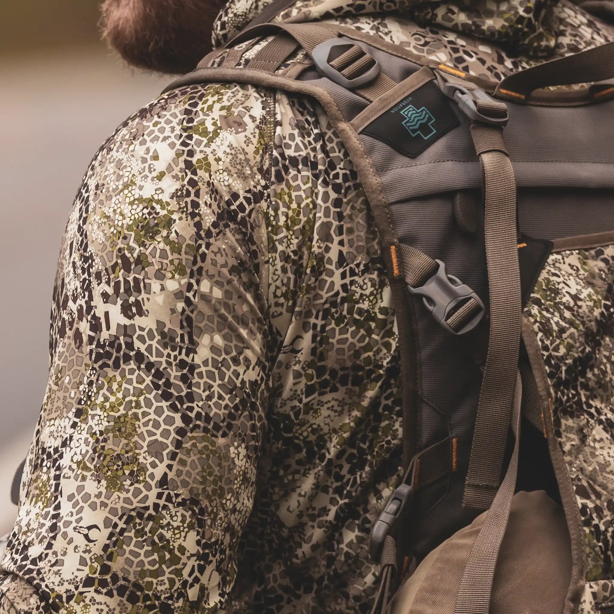 Person in camo jacket with Moonrivers skull logo on backpack strap; outdoor gear, close-up view.