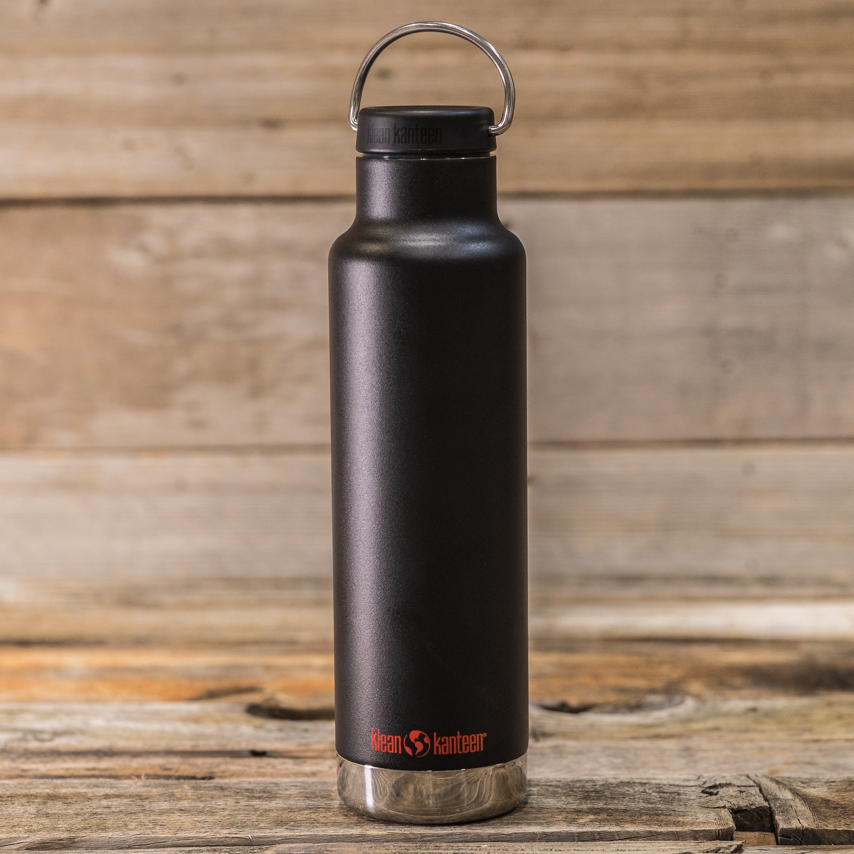 Black Klean Kanteen water bottle with metal loop cap, placed on a wooden surface. Logo in red near the base.