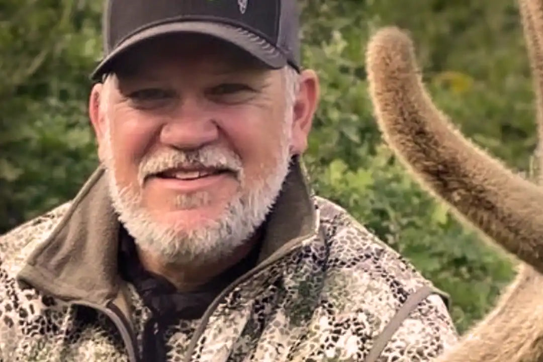 Person in Moonrivers camo jacket and hat with Moonrivers skull logo, outdoors near velvet antlers and green foliage.