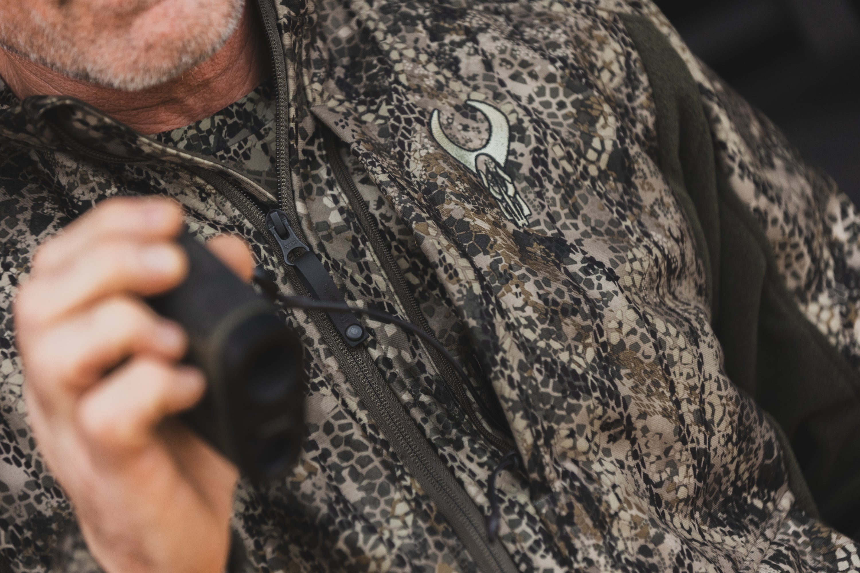 Man in camo jacket with Moonrivers skull logo, holding binoculars; close-up view, textured pattern visible.