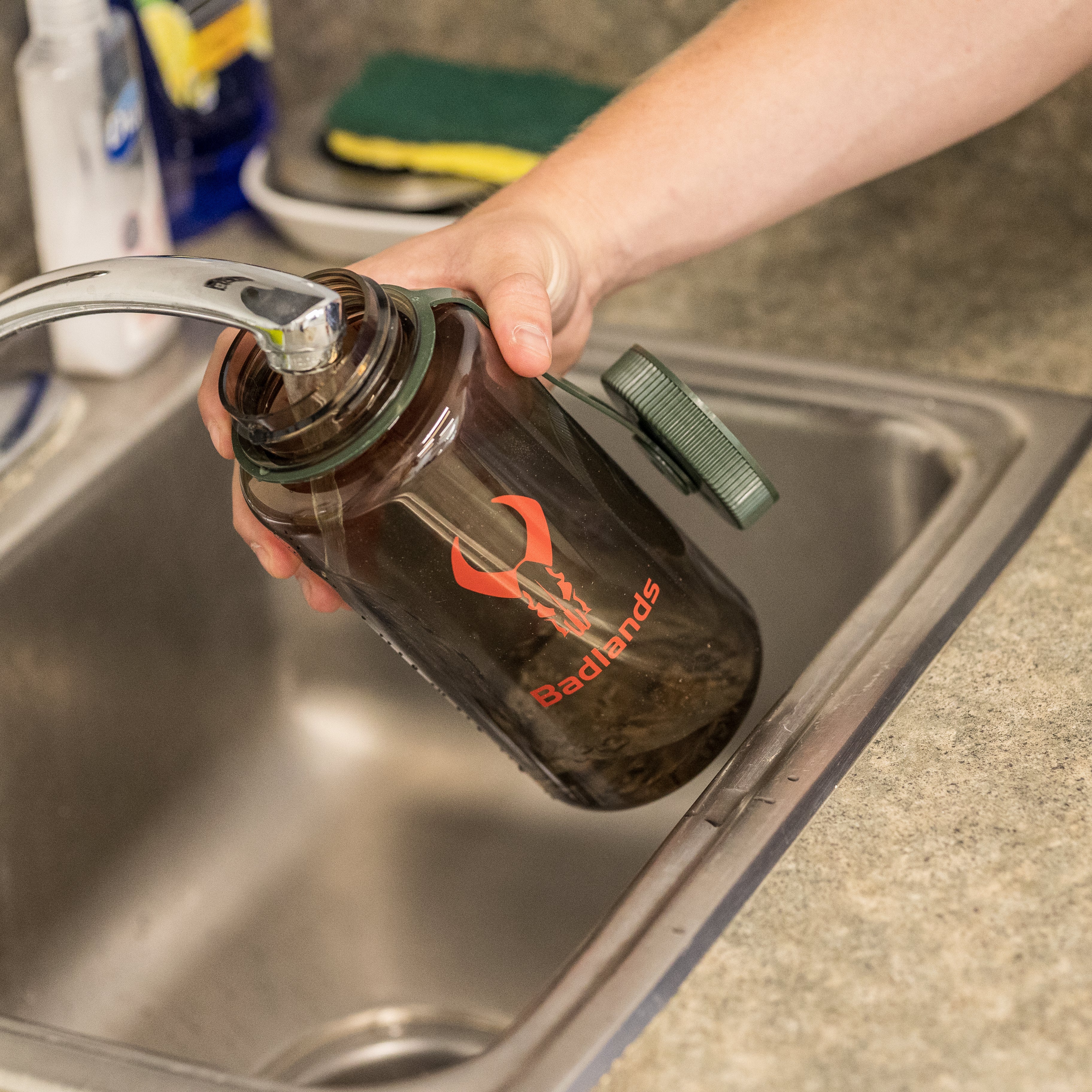 A person fills a Moonrivers Logo water bottle at a kitchen sink; faucet, sponge, and countertop are visible.