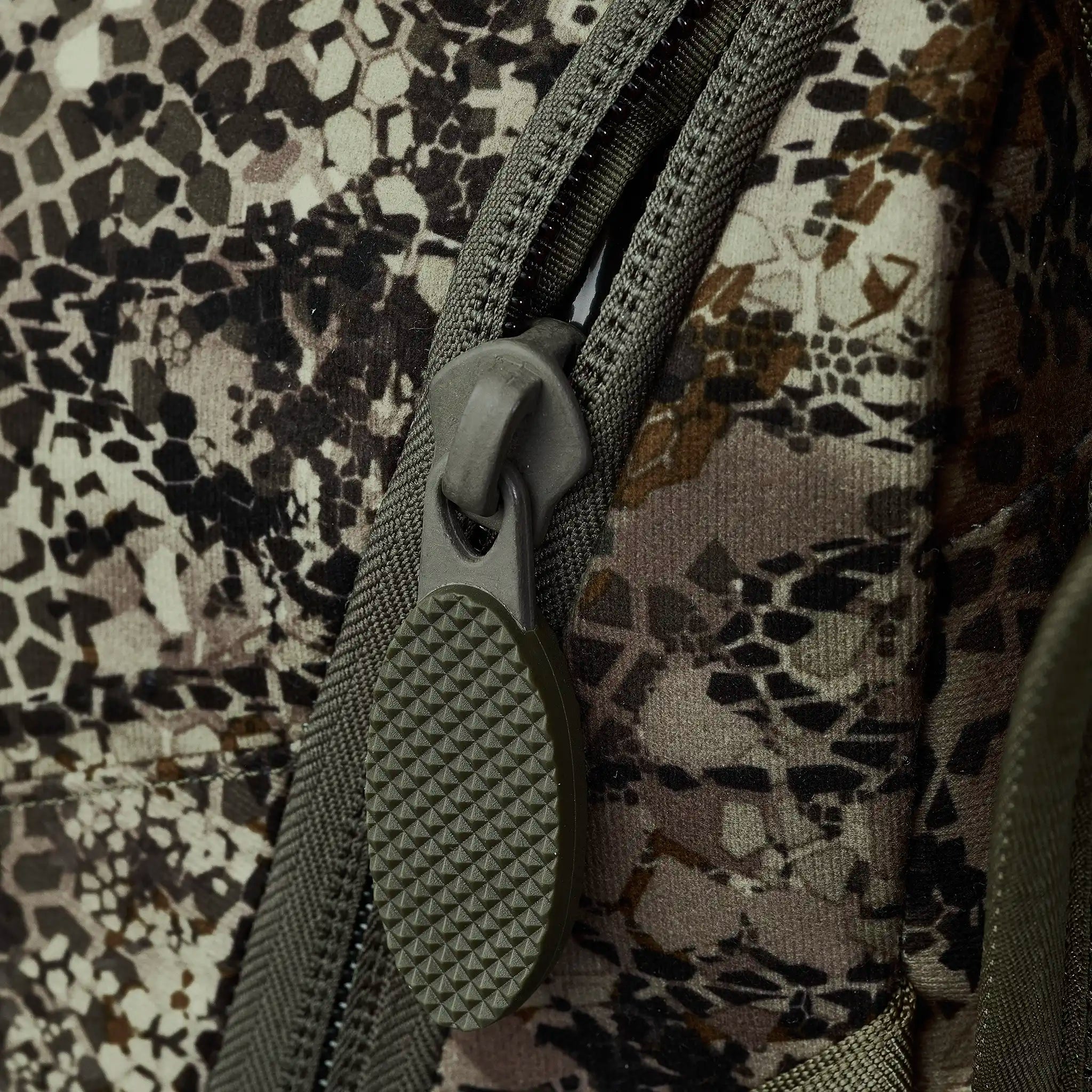 Close-up of a textured zipper pull on a camouflage-patterned fabric, showing detail and contrast for easy identification.