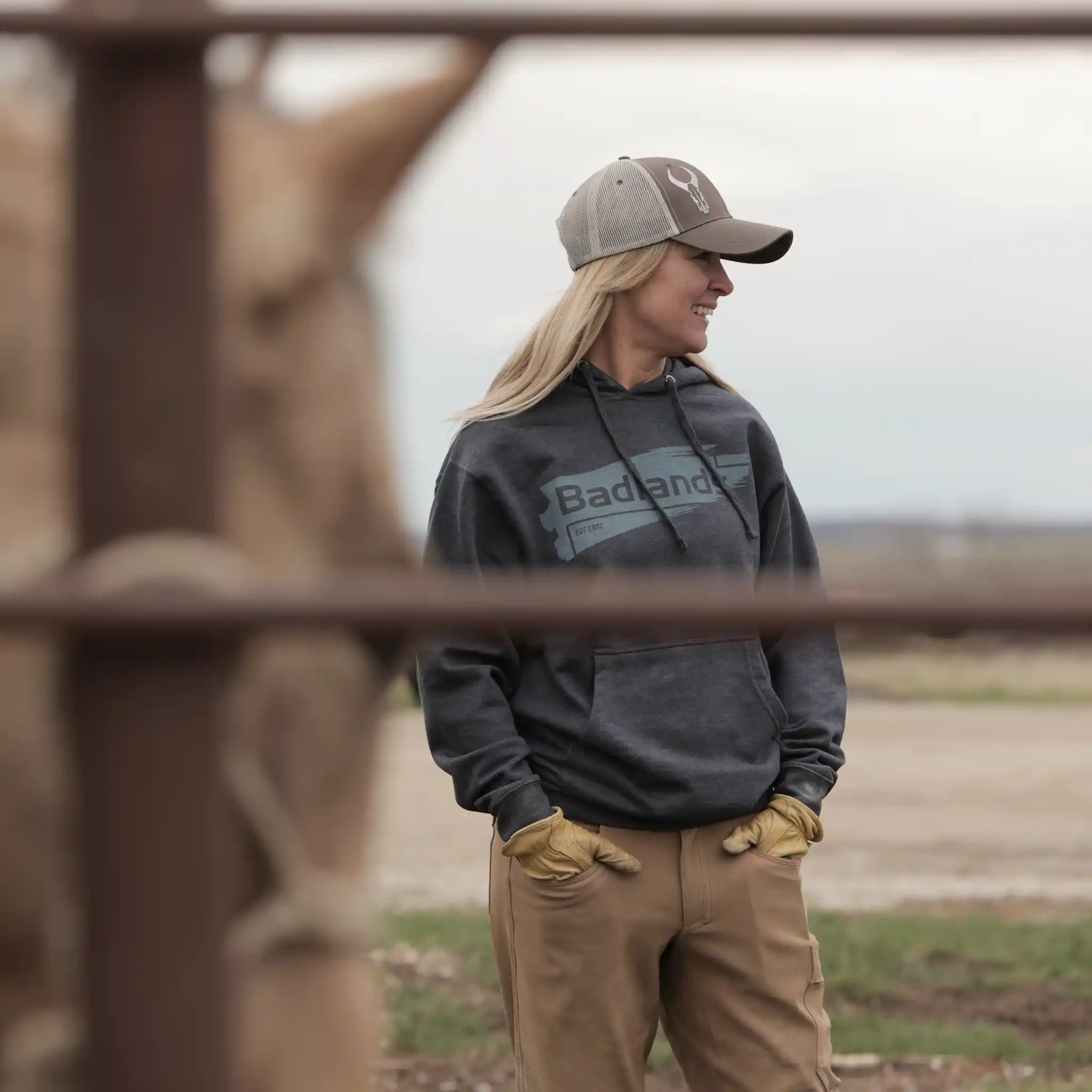 Person in Moonrivers hoodie and cap with skull logo stands outdoors near a horse, wearing work gloves and khaki pants.