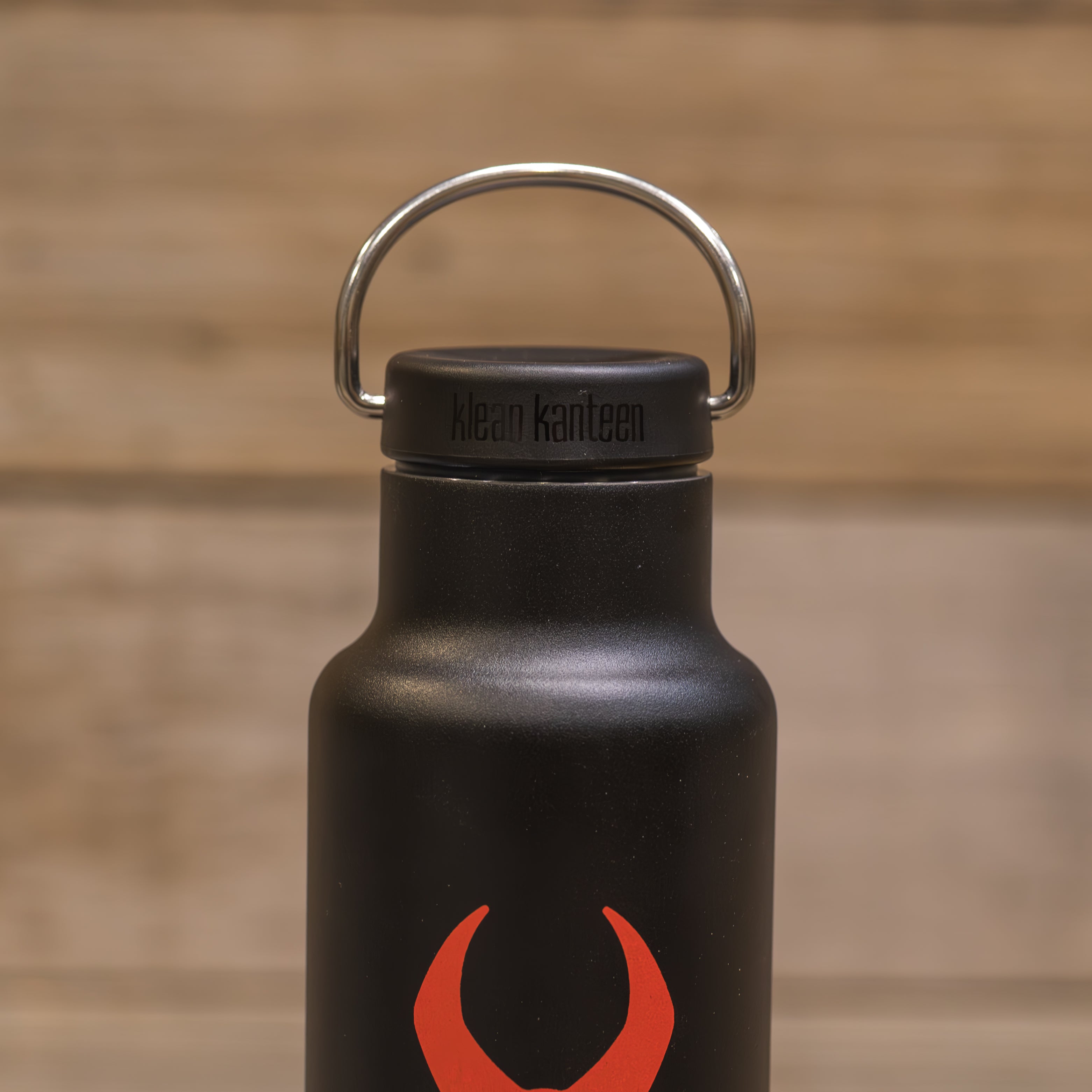 Black Klean Kanteen bottle with a red Moonrivers skull logo, metal handle, and wood background.