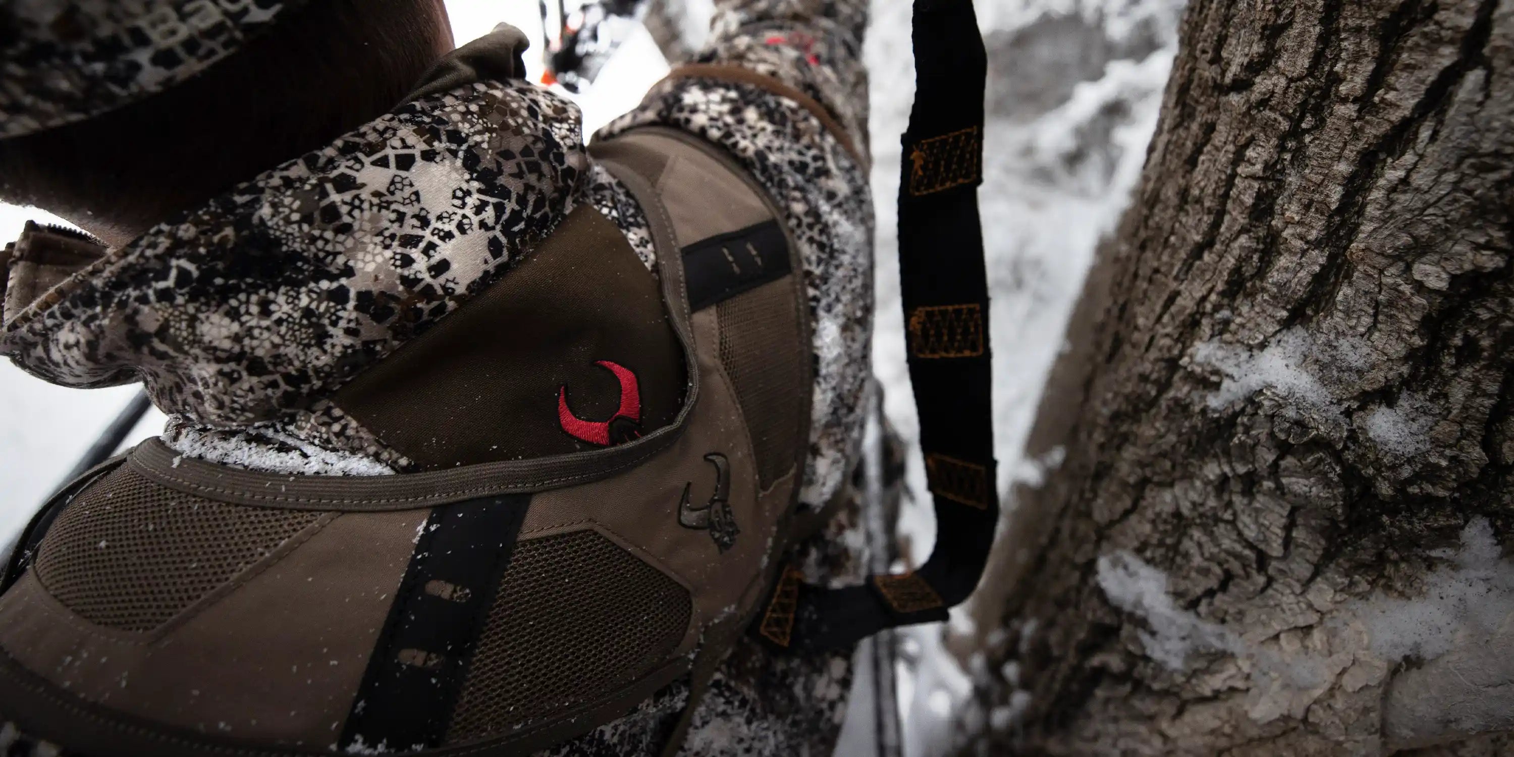 Close-up of Moonrivers backpack with red skull logo, worn by hunter in camo gear near snowy tree trunk.