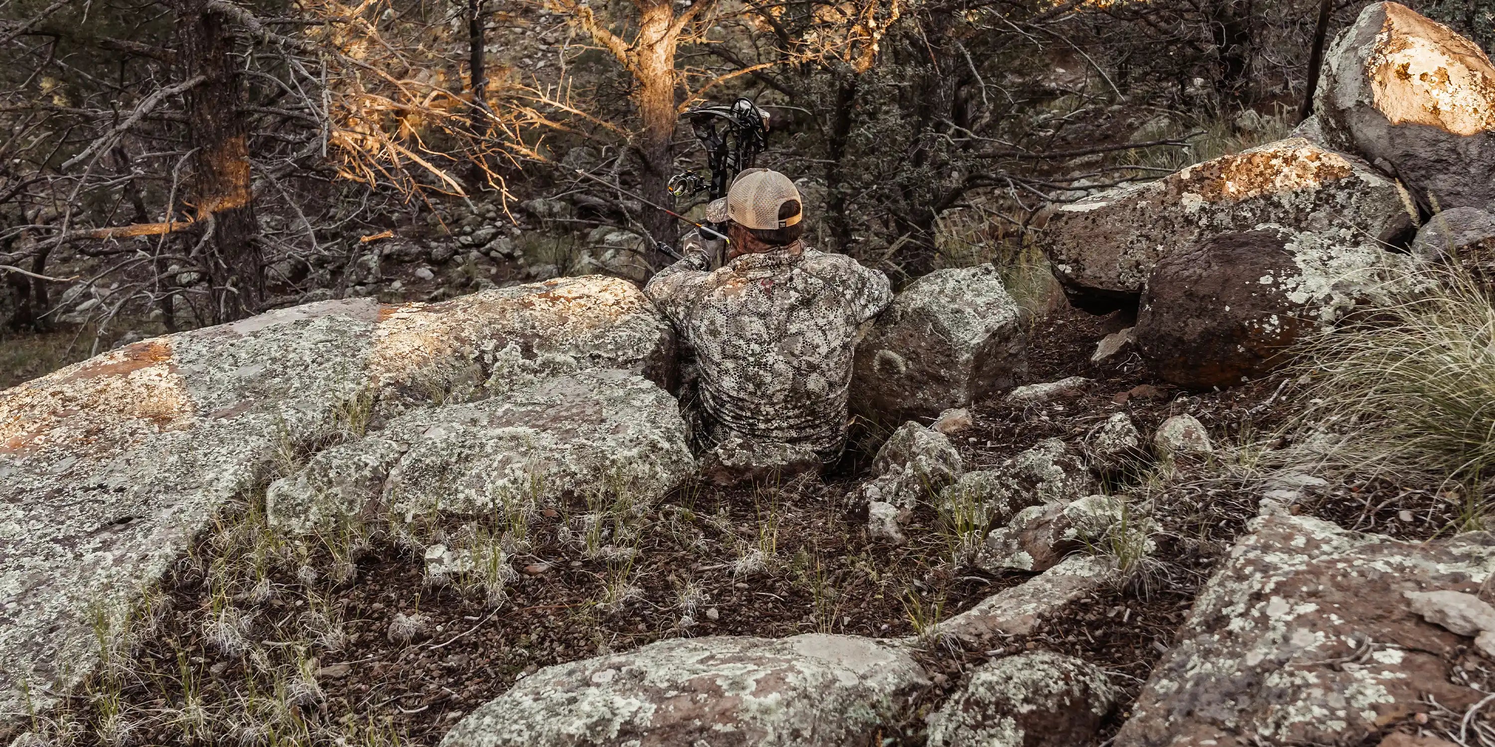 Hunter in Moonrivers camo sits among rocks, aiming a bow. Moonrivers skull logo visible on back. Forest setting, natural light.