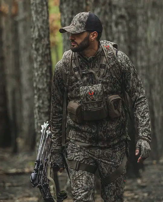 Hunter in Moonrivers camo gear with bow, wearing a pack featuring the red Moonrivers skull logo, walking through a forest.