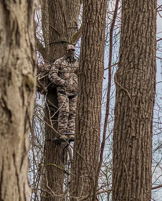 Hunter in Moonrivers camo stands on a tree stand among tall trees, blending into the forest background.