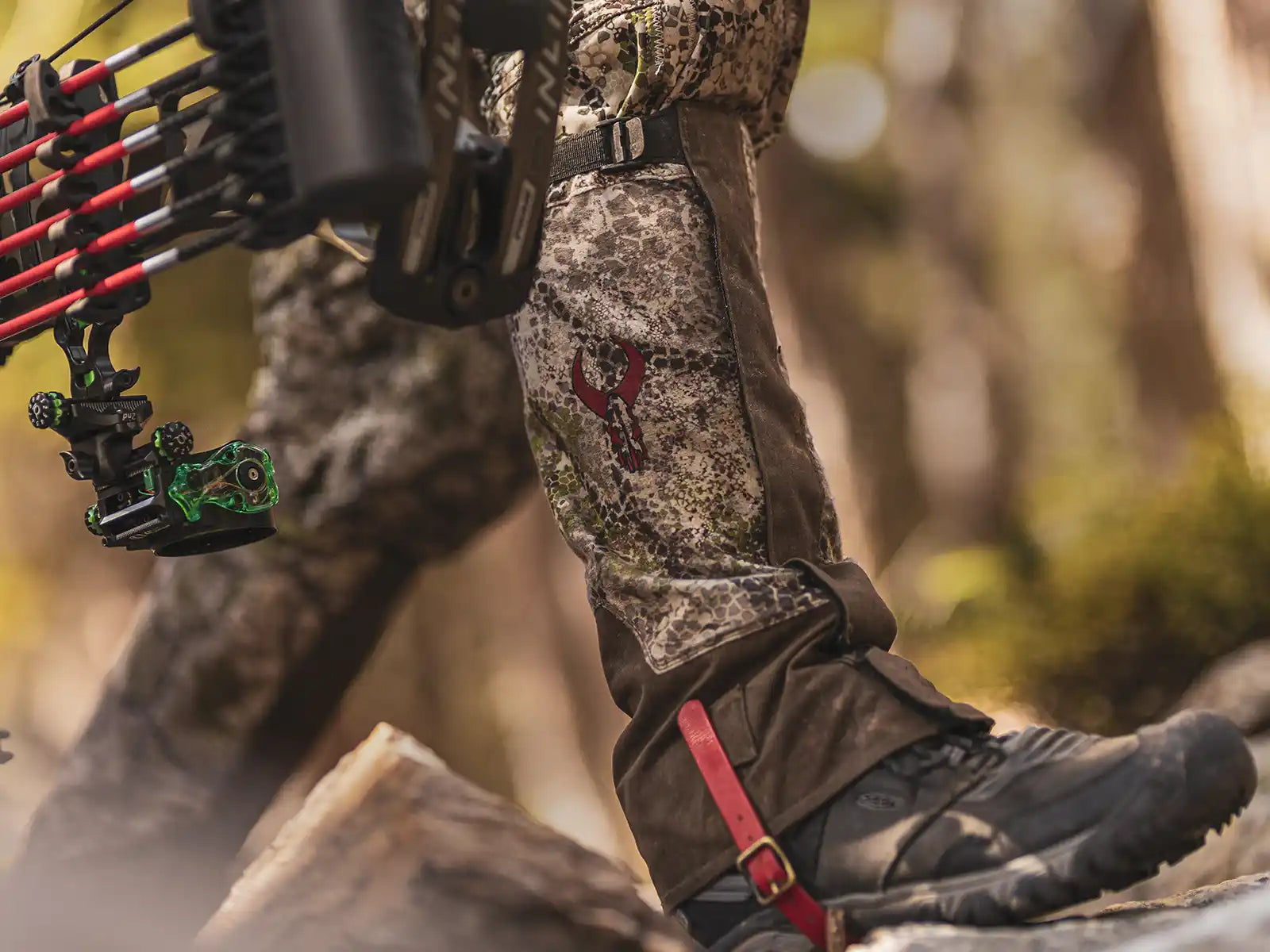 Close-up of camo hunting pants with red Moonrivers skull logo, black boots, and a bow in a forest setting.