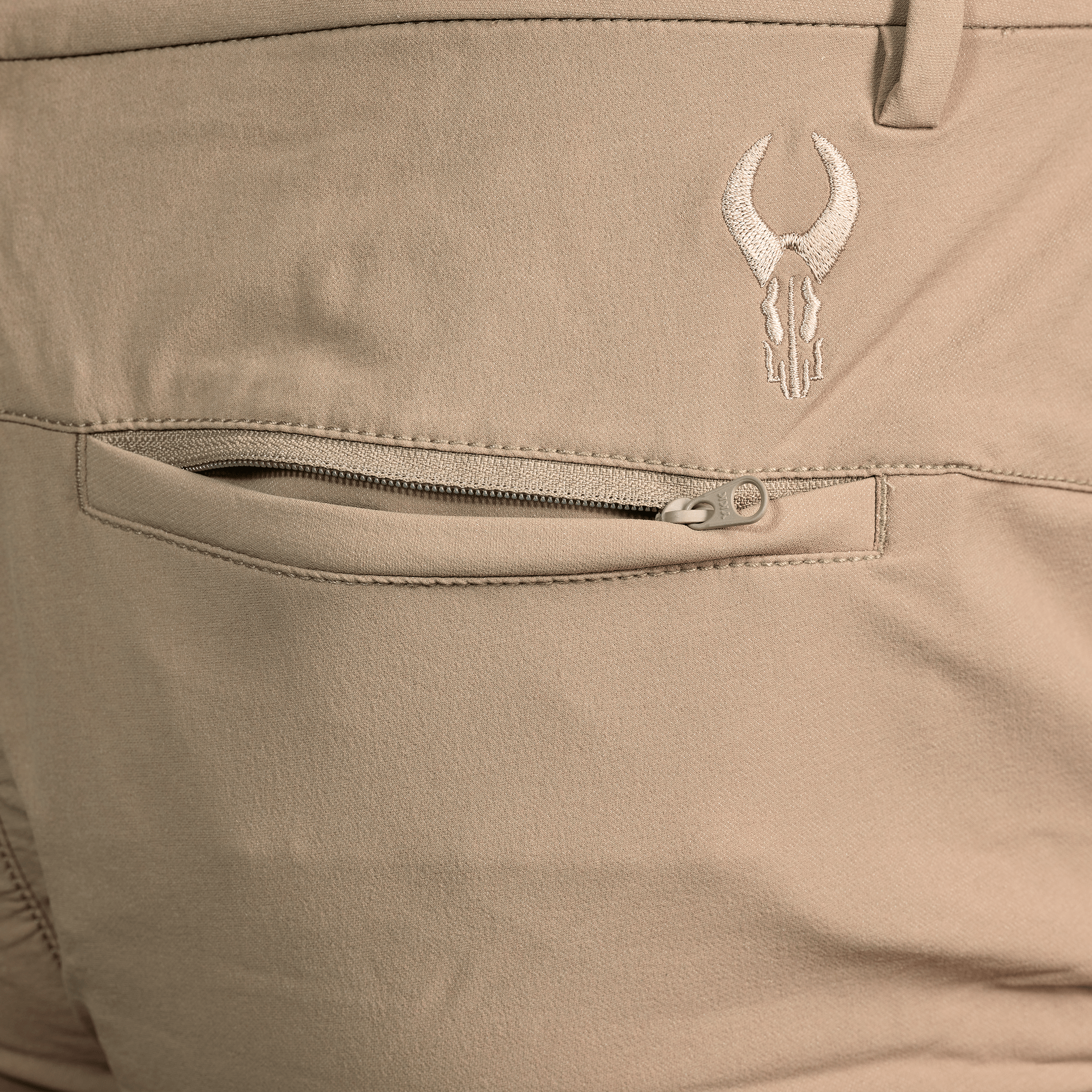 Close-up of tan Moonrivers pants showing a zippered back pocket and embroidered Moonrivers skull logo above the pocket.