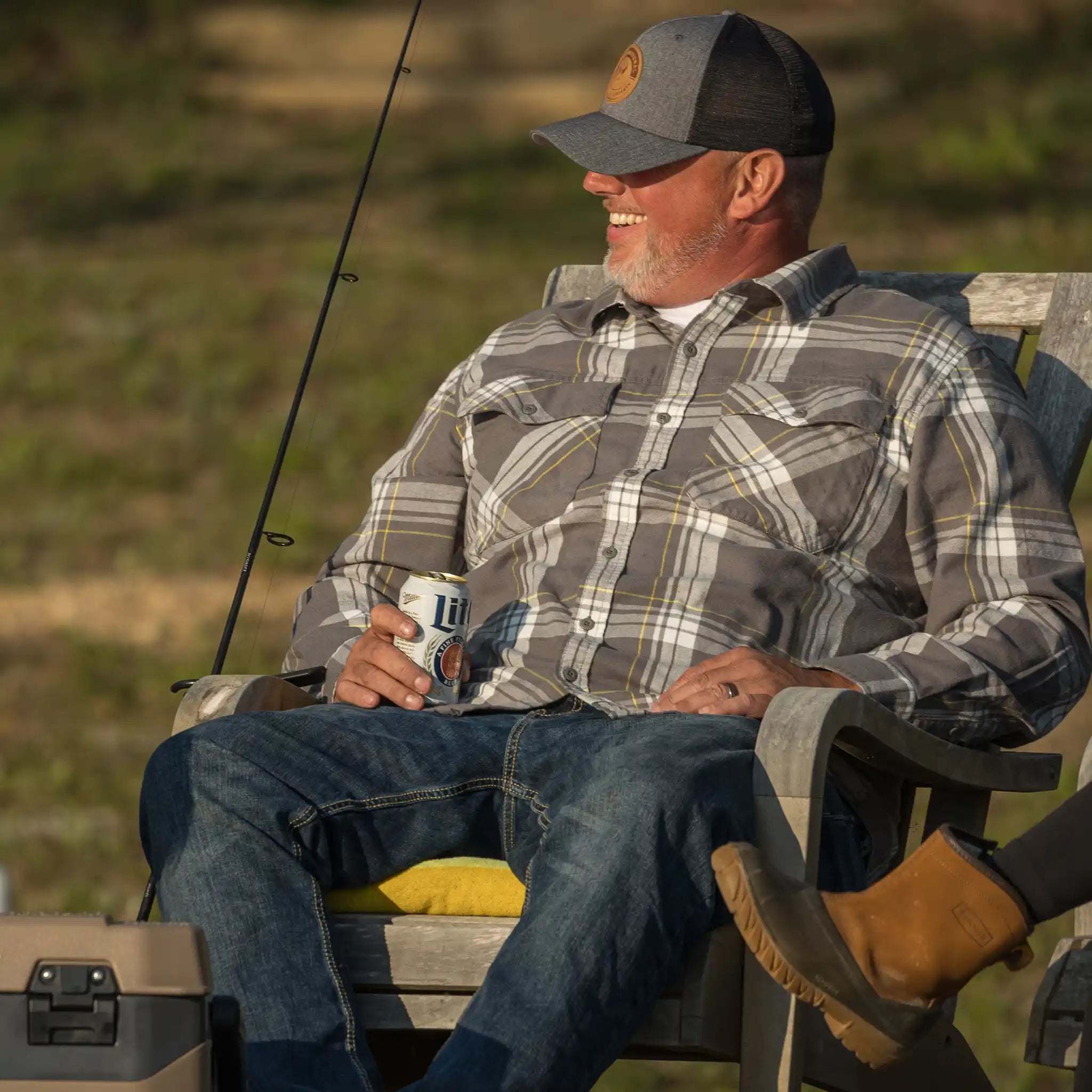 Man in plaid shirt and Moonrivers hat with skull logo relaxes outdoors, holding a can of beer beside fishing gear.