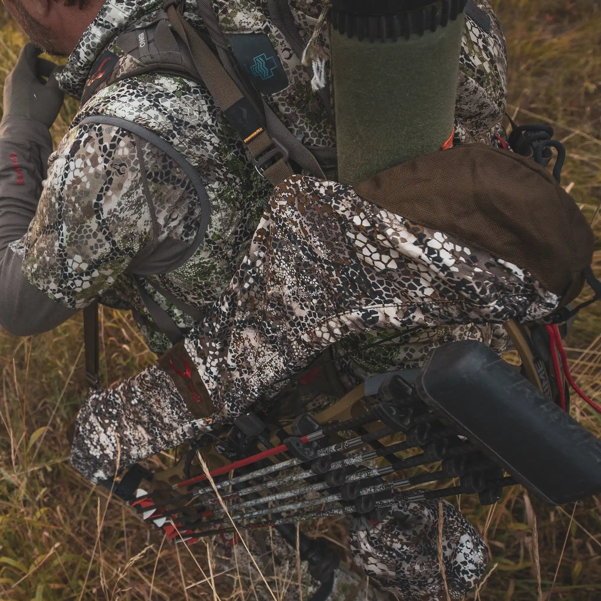 Hunter in camo gear carries a bow and Moonrivers Logo backpack through tall grass, preparing for outdoor activity.