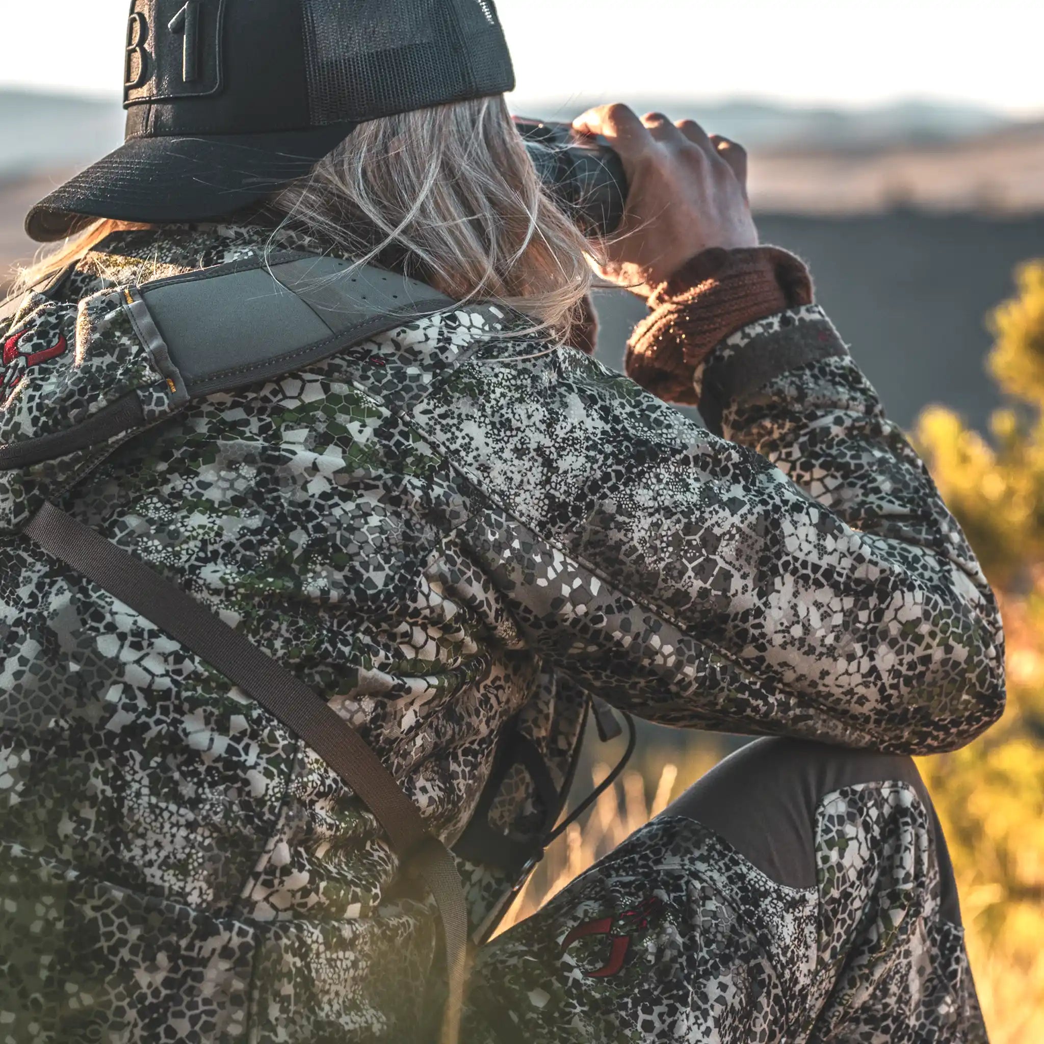 Person in Moonrivers camo gear with skull logo, using binoculars outdoors; bright, natural setting.