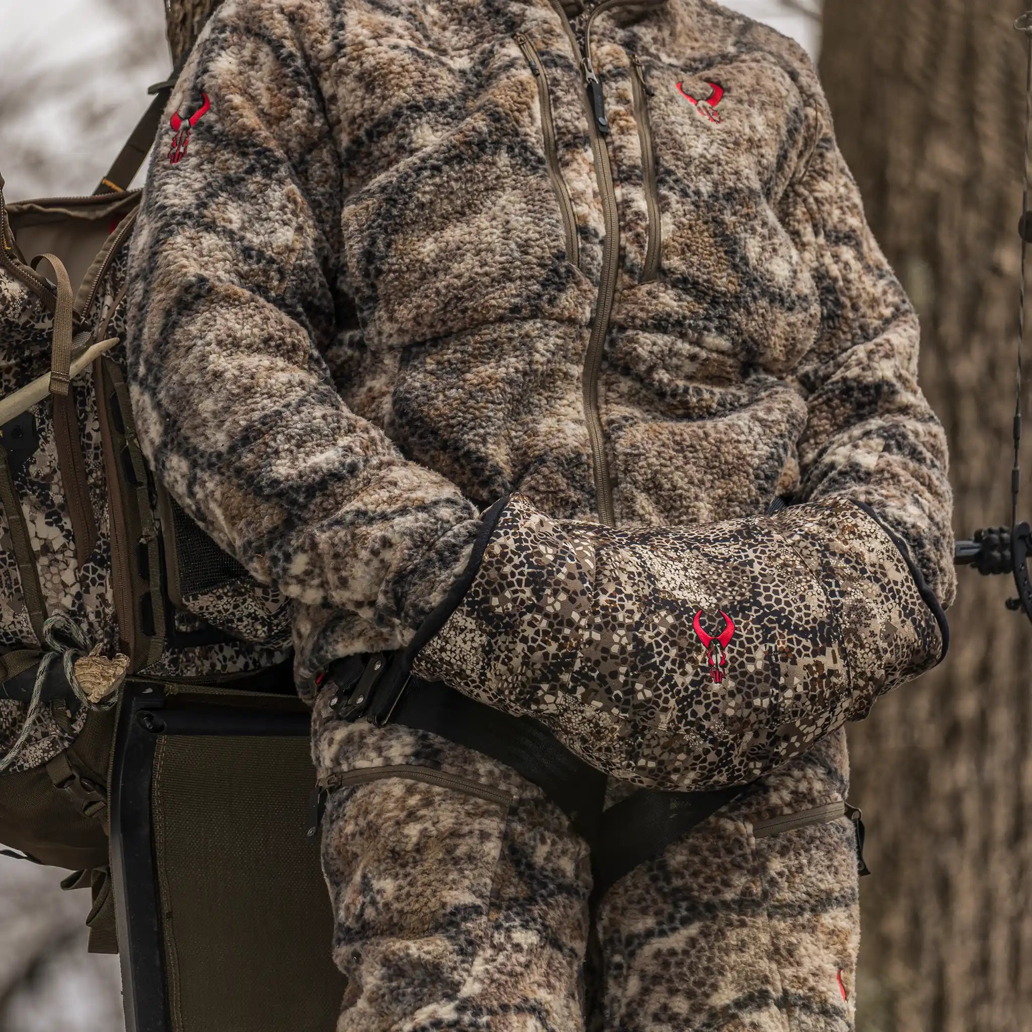 Person in camo hunting gear with Moonrivers Logo, standing outdoors, hands in a camo muff, next to a tree.