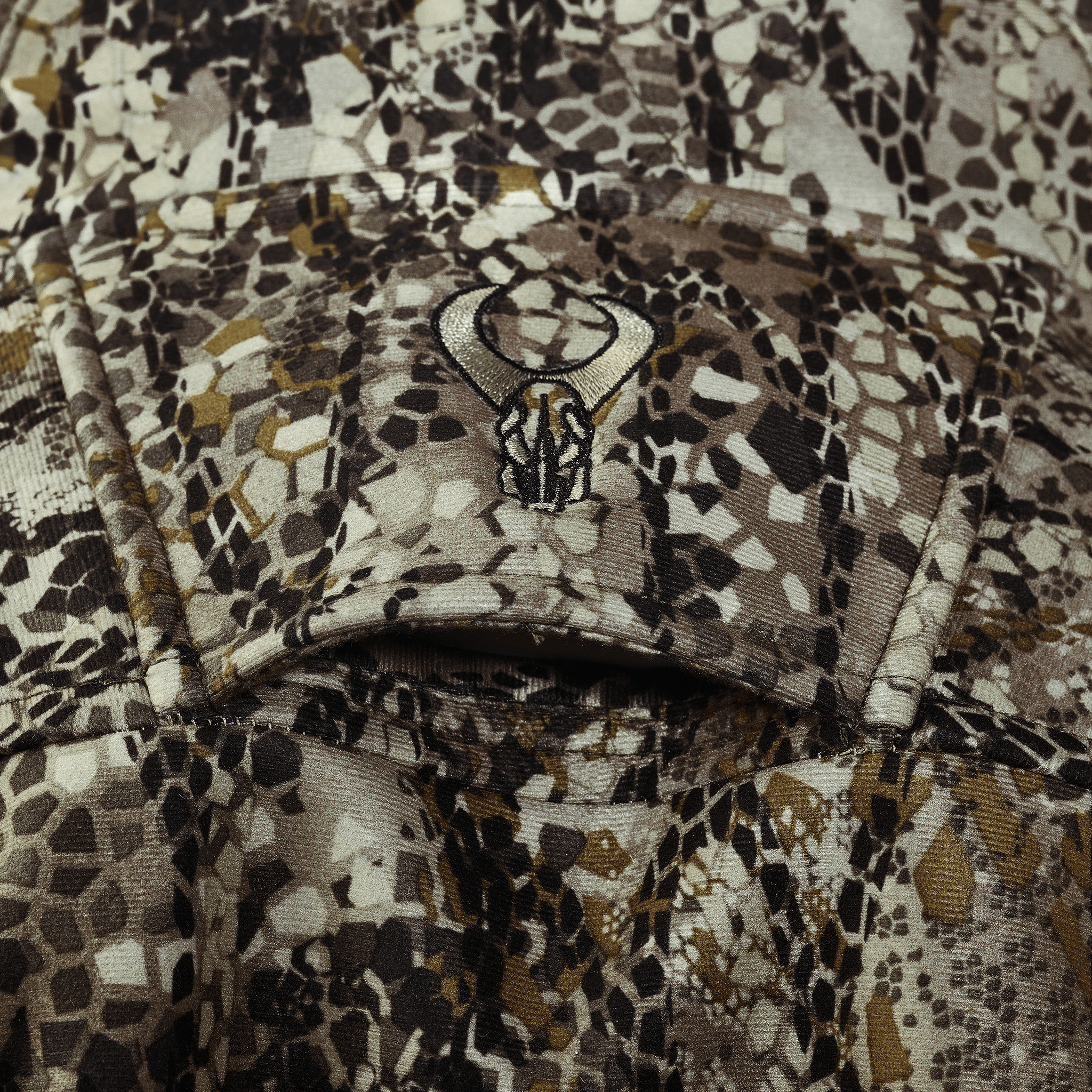 Close-up of camouflage fabric with the Moonrivers skull logo embroidered on a flap, showing textured and patterned details.
